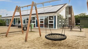 Un parque infantil con columpios y un neumático colgante, un edificio con ventanales grandes y una mesa de picnic.