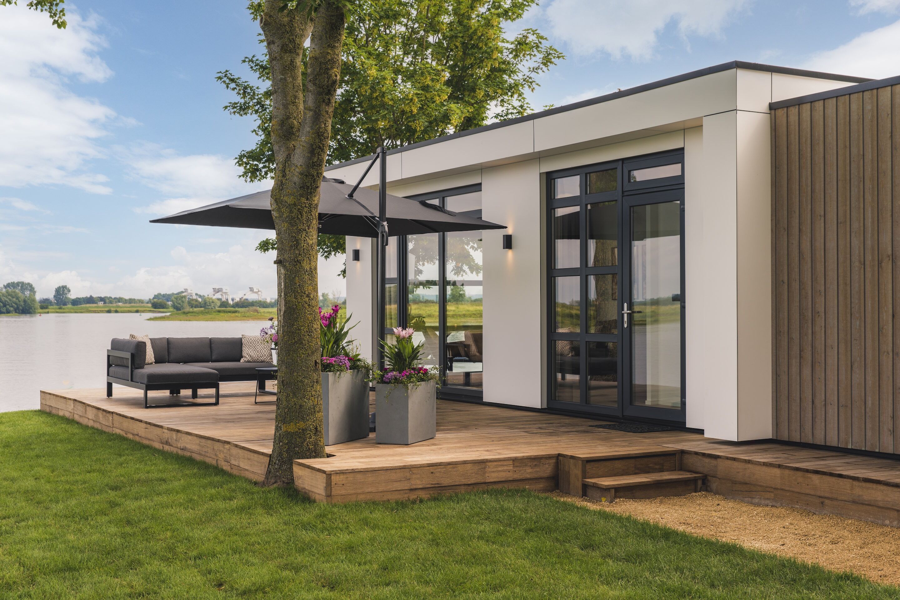 Una casa moderna con terraza, muebles de exterior y un gran ventanal con vistas a un lago.
