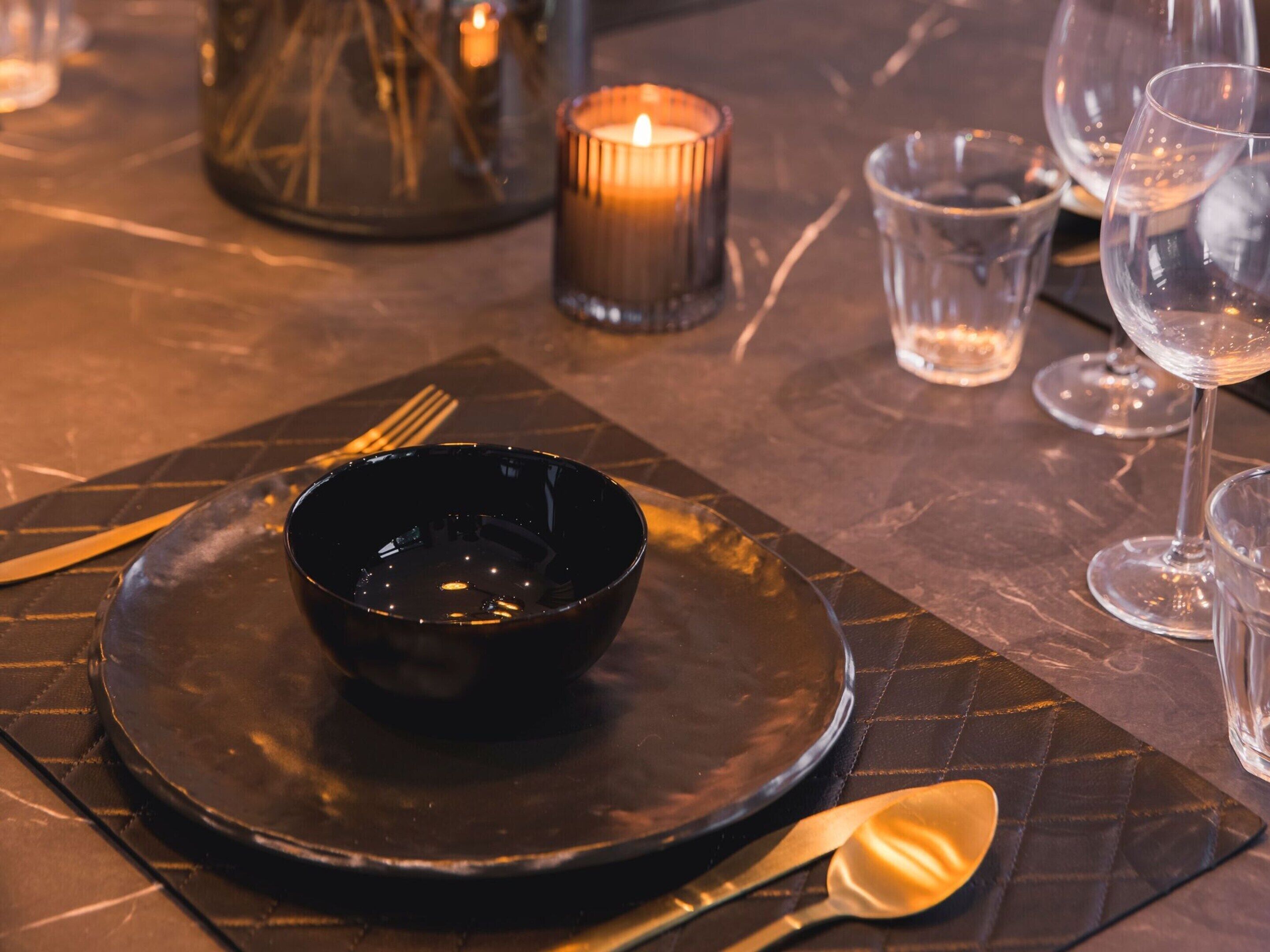 A table set with a black bowl, cutlery, and wine glasses, with a lit candle in the background.