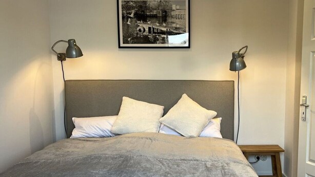 Una cama bien hecha con cabecera gris, dos lámparas de noche y una fotografía en blanco y negro enmarcada en la pared.
