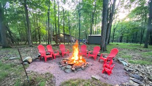 A campfire with red Adirondack chairs arranged around it in a forested area. - Eat, Sleep, Ski. Repeat.  (Lake Harmony)