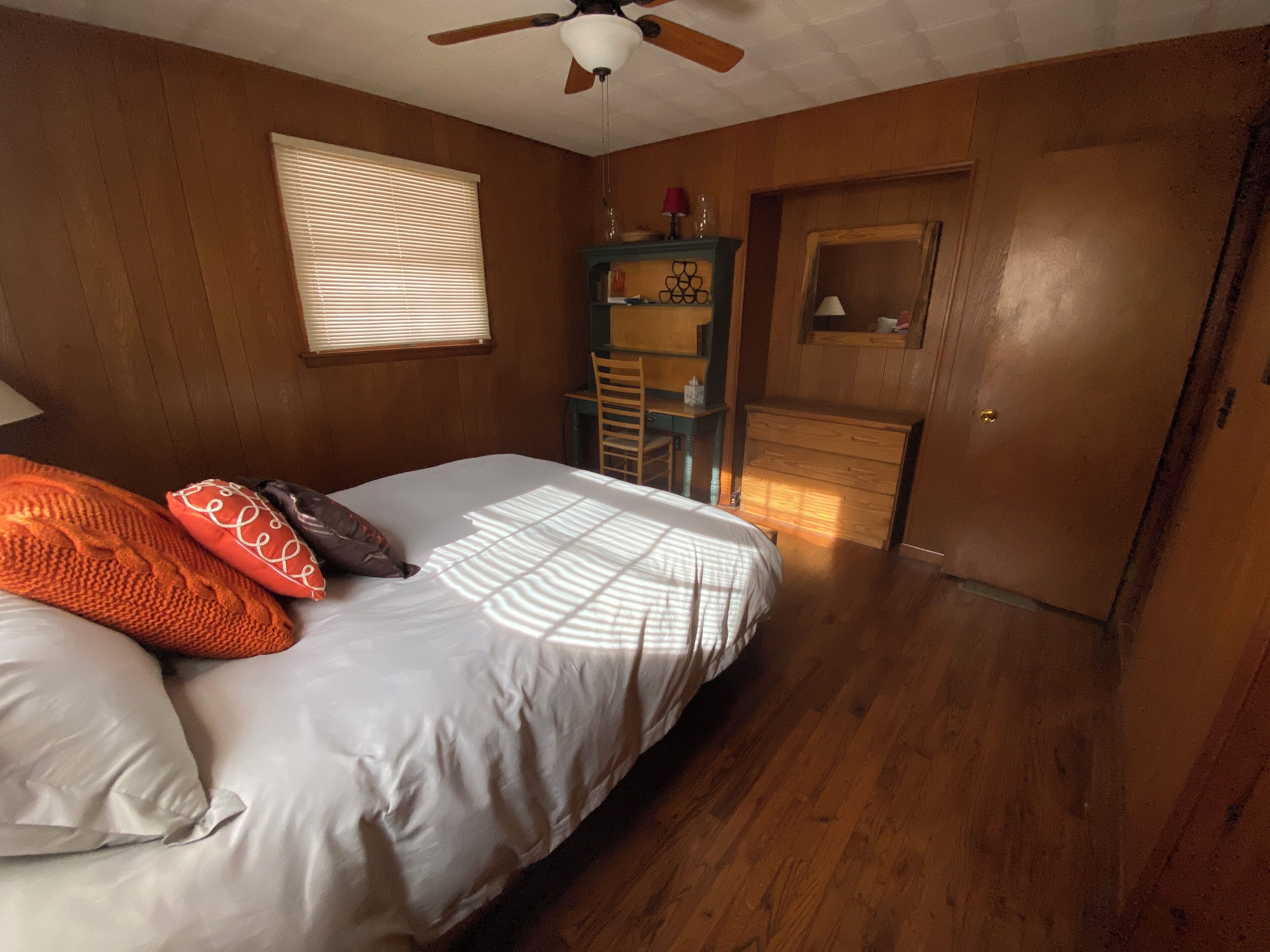 Une chambre à coucher avec un lit, des murs en bois, un ventilateur de plafond, une fenêtre avec des stores, une commode et un miroir.