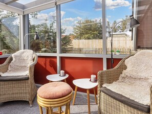 Una habitación iluminada por el sol, con muebles de mimbre, una mesita pequeña y vistas a una valla de madera a través de amplios ventanales.