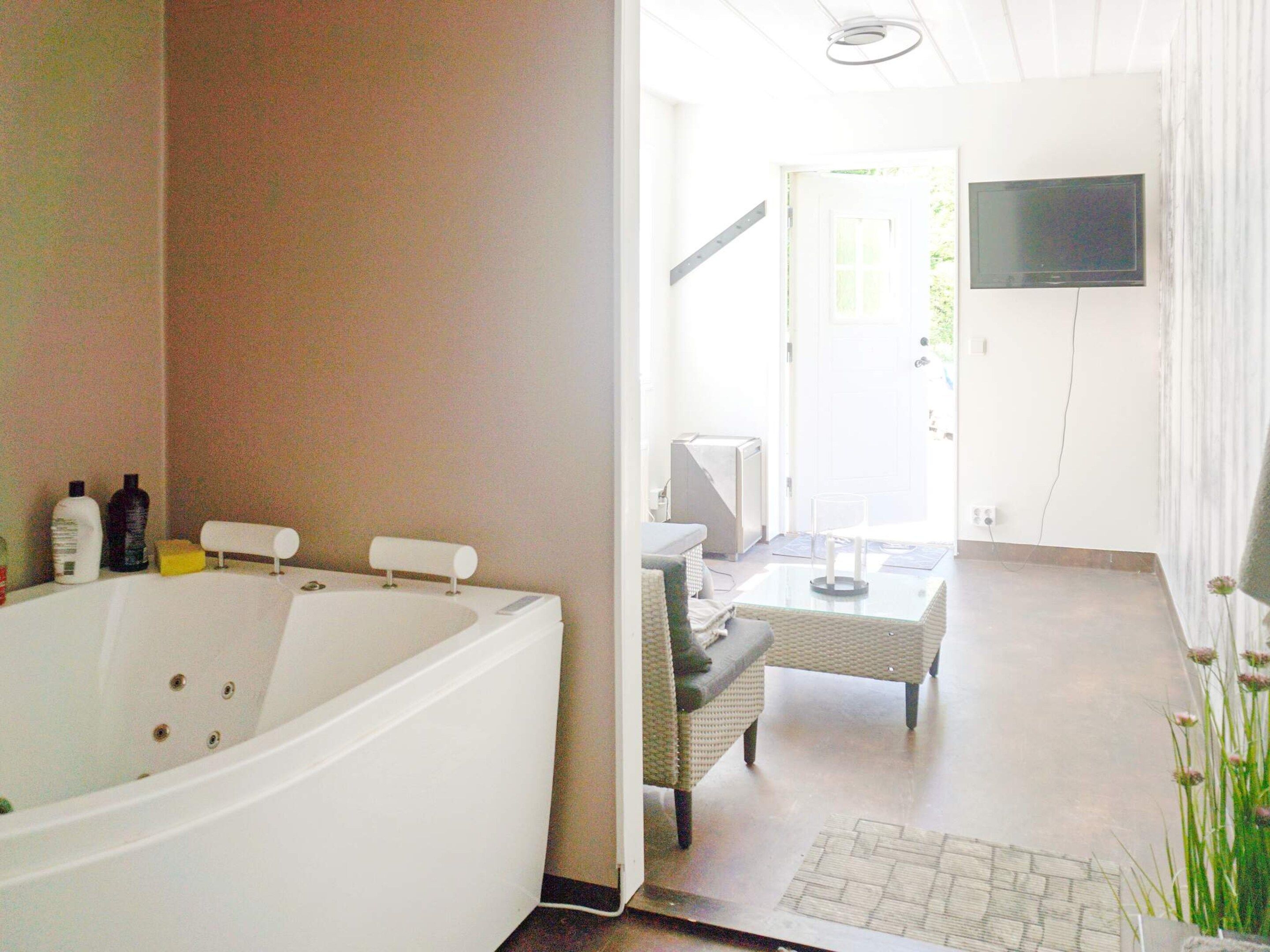 A bathroom with a large bathtub, a sink, and toiletries. There is a television mounted on the wall and a wicker chair in the adjacent living area.