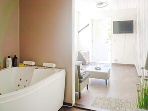 A bathroom with a large bathtub, a sink, and toiletries. There is a television mounted on the wall and a wicker chair in the adjacent living area. - 4 Star Holiday Home in Hallnas (Hållnäs)