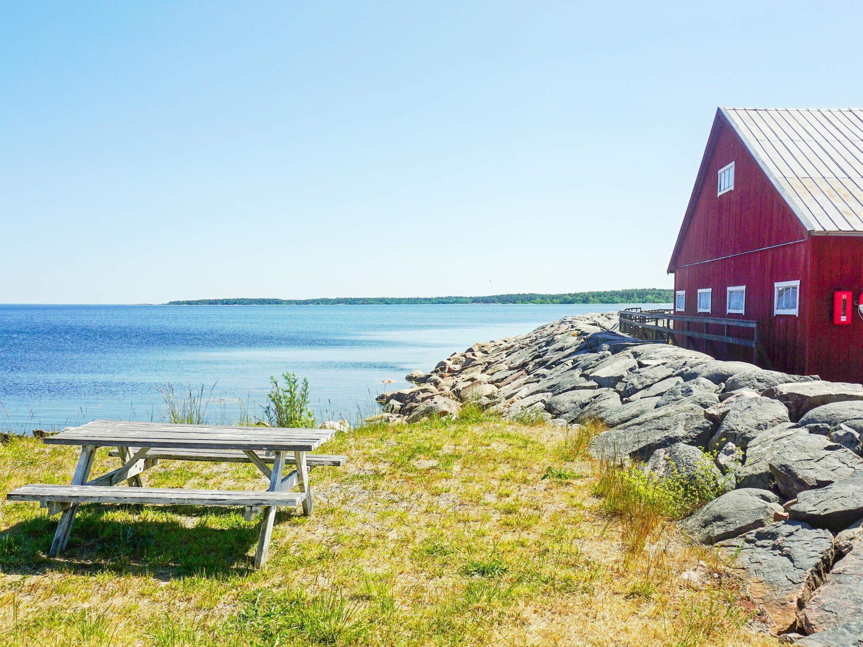 A red wooden building with a dock extending into a calm body of water, a picnic table in the foreground, and a rocky shoreline.