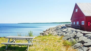 A red wooden building with a dock extending into a calm body of water, a picnic table in the foreground, and a rocky shoreline.