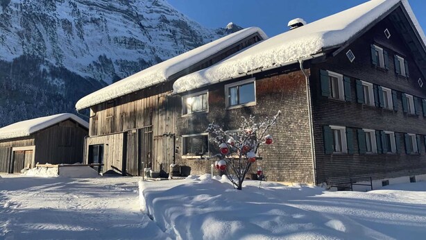 A wooden building with a snow-covered roof, a brick section, and a small tree with red ornaments in front. - Holiday Home Geser Comfortable Vacation Home (Schnepfau)