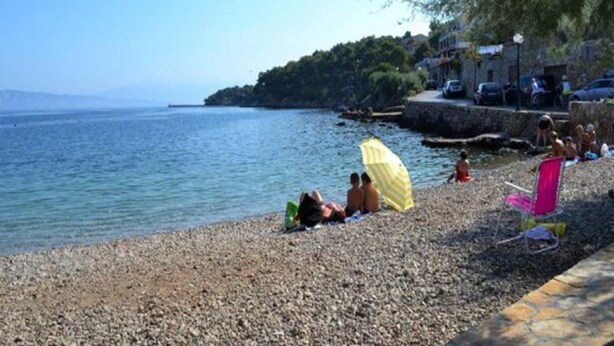 Una playa pedregosa con gente tomando el sol y un paraguas amarillo.