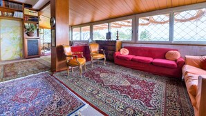 A cozy living room with a red sofa, two armchairs, and a coffee table. There is a bookshelf filled with books and a potted plant. The room has large windows with a view of greenery outside. - Belvilla by OYO Villa Teodora (Anguillara Sabazia)