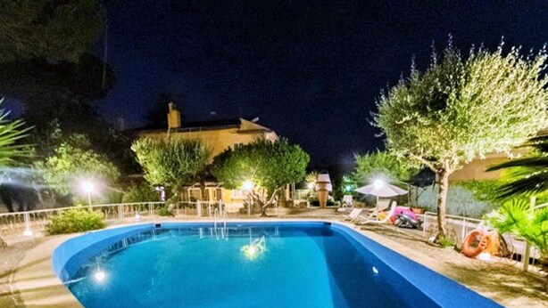 A nighttime swimming pool with a curved edge, surrounded by a low fence and various trees. - Belvilla by OYO Villa Teodora (Anguillara Sabazia)