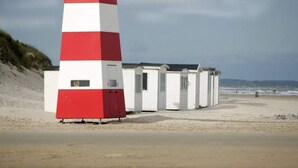 Un faro rosso e bianco accanto a una fila di casette bianche sulla spiaggia, con una spiaggia sabbiosa e un cielo nuvoloso sullo sfondo.