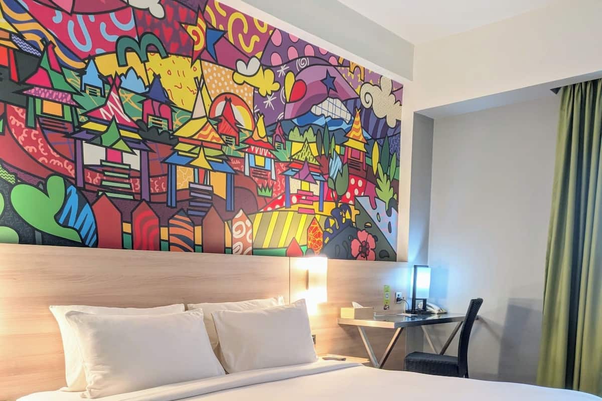 Hotellihuone, jossa on suuri sänky, yöpöytä, työpöytä lampun kanssa ja värikäs abstrakti seinämaalaus.