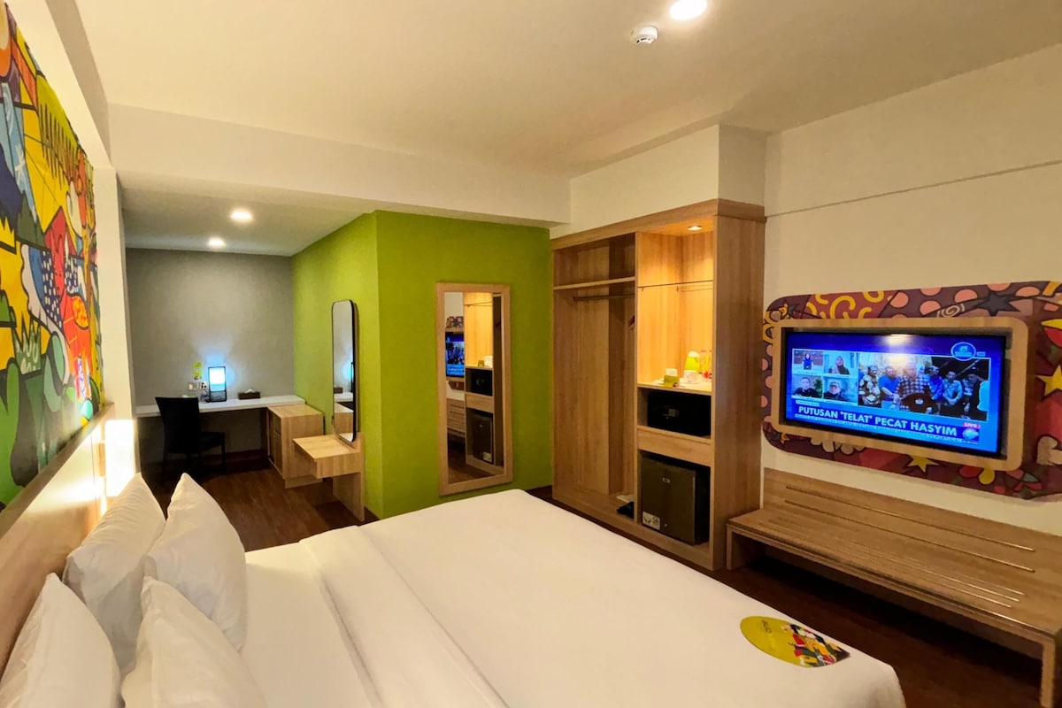 A hotel room with a bed, a desk, a television, and a colorful mural on the wall.