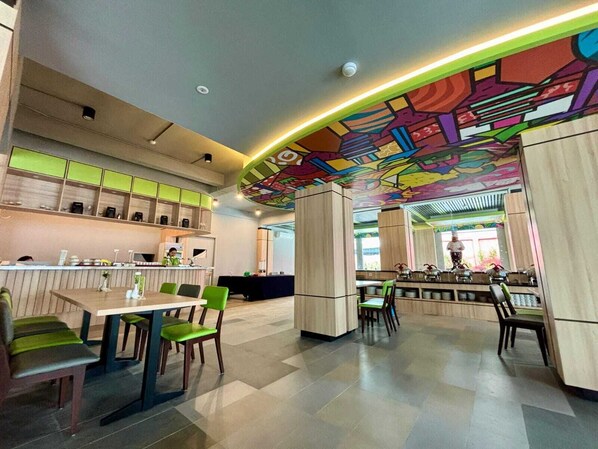En moderne restaurant med et farverigt muralmaleri i loftet, et barområde og siddepladser med grønne stole.