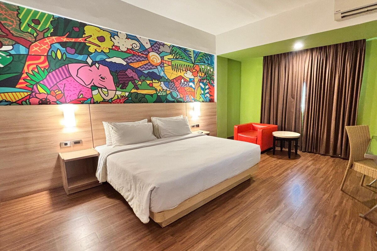 Habitación de hotel con una cama grande, un mural colorido en la pared, una pared de acento verde, una silla roja, una pequeña mesa redonda y un mueble de televisión de vidrio.