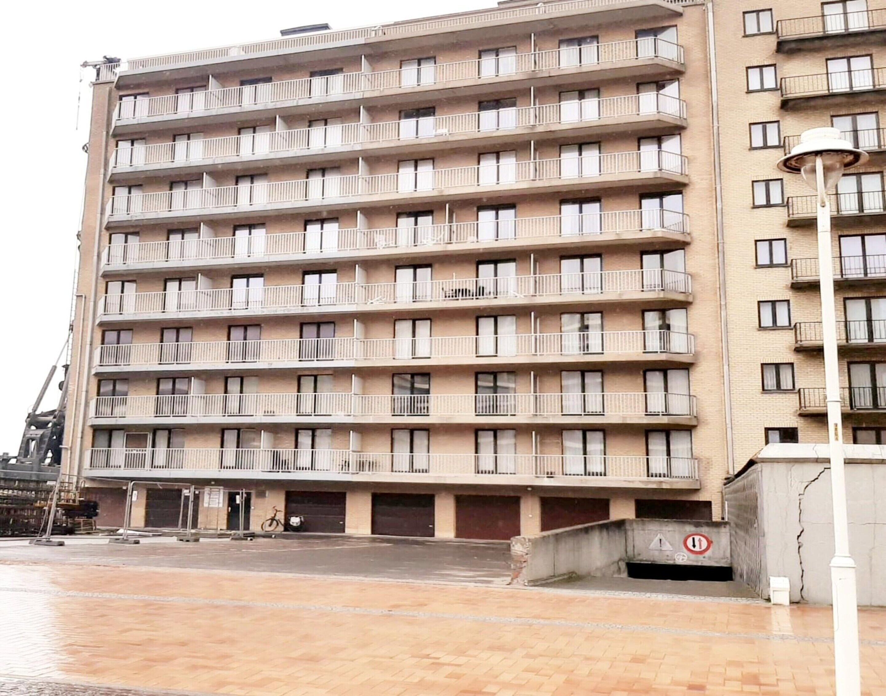 A multi-story residential building with balconies, a parking area, and a pedestrian walkway.