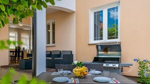 An outdoor patio with a dining table set for four, surrounded by wicker chairs. A grill is visible in the background, and there is a potted plant with blue flowers to the right. - Andrea Comfortable Holiday Residence (Marcana)
