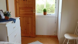 Ein Schlafzimmer mit Holztür, einer weißen Kommode, einem Stuhl, einem Bett mit gemustertem Überwurf und einem kleinen Topfpflanzen auf der Fensterbank. Durch das Fenster ist Grünlandschaft sichtbar.