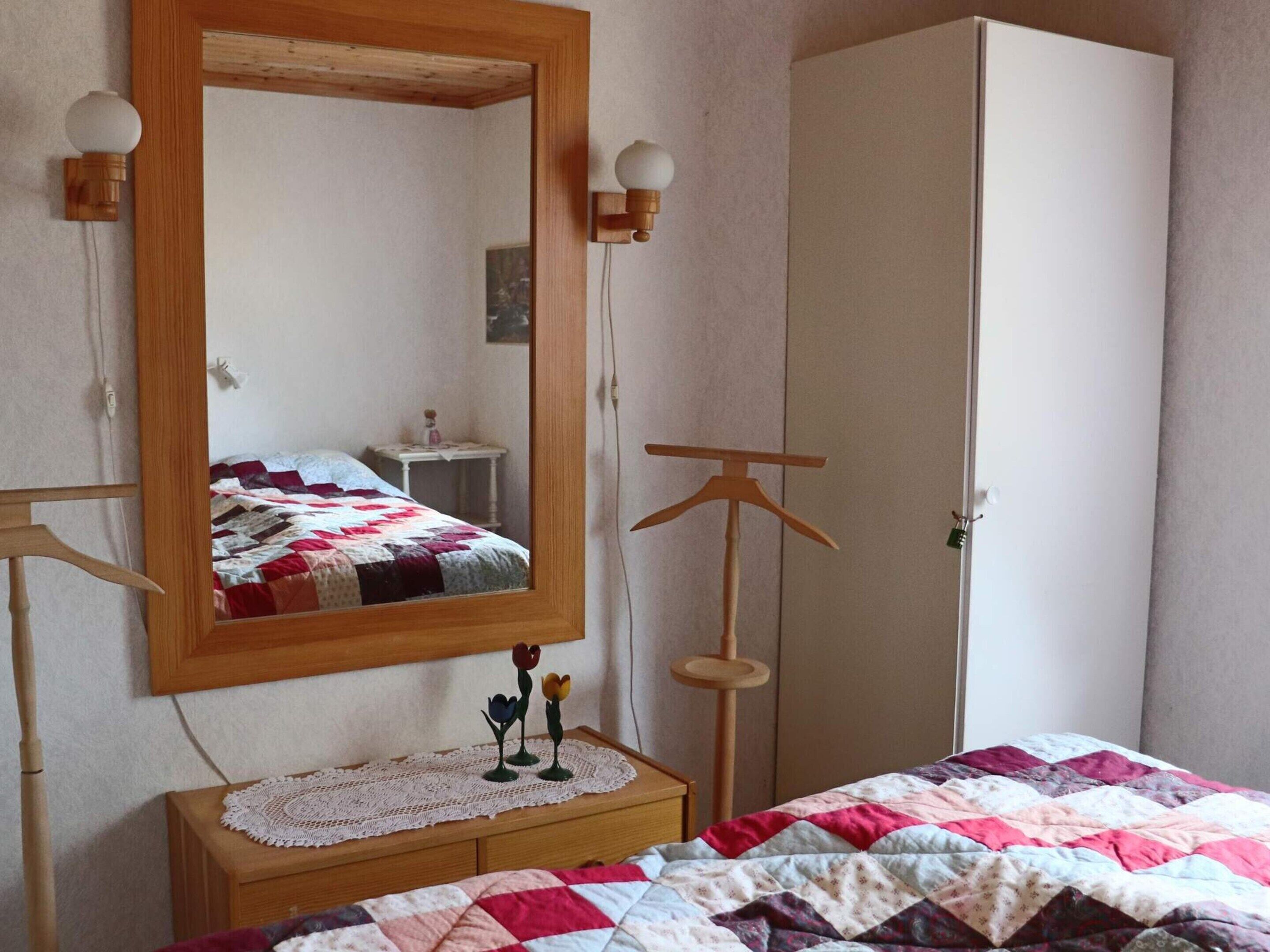 Ein Schlafzimmer mit einem Holfischschrank, einem Nachttisch mit Spitzendecke, einem Spiegel und einem Bett mit gesteckter Bettdecke.
