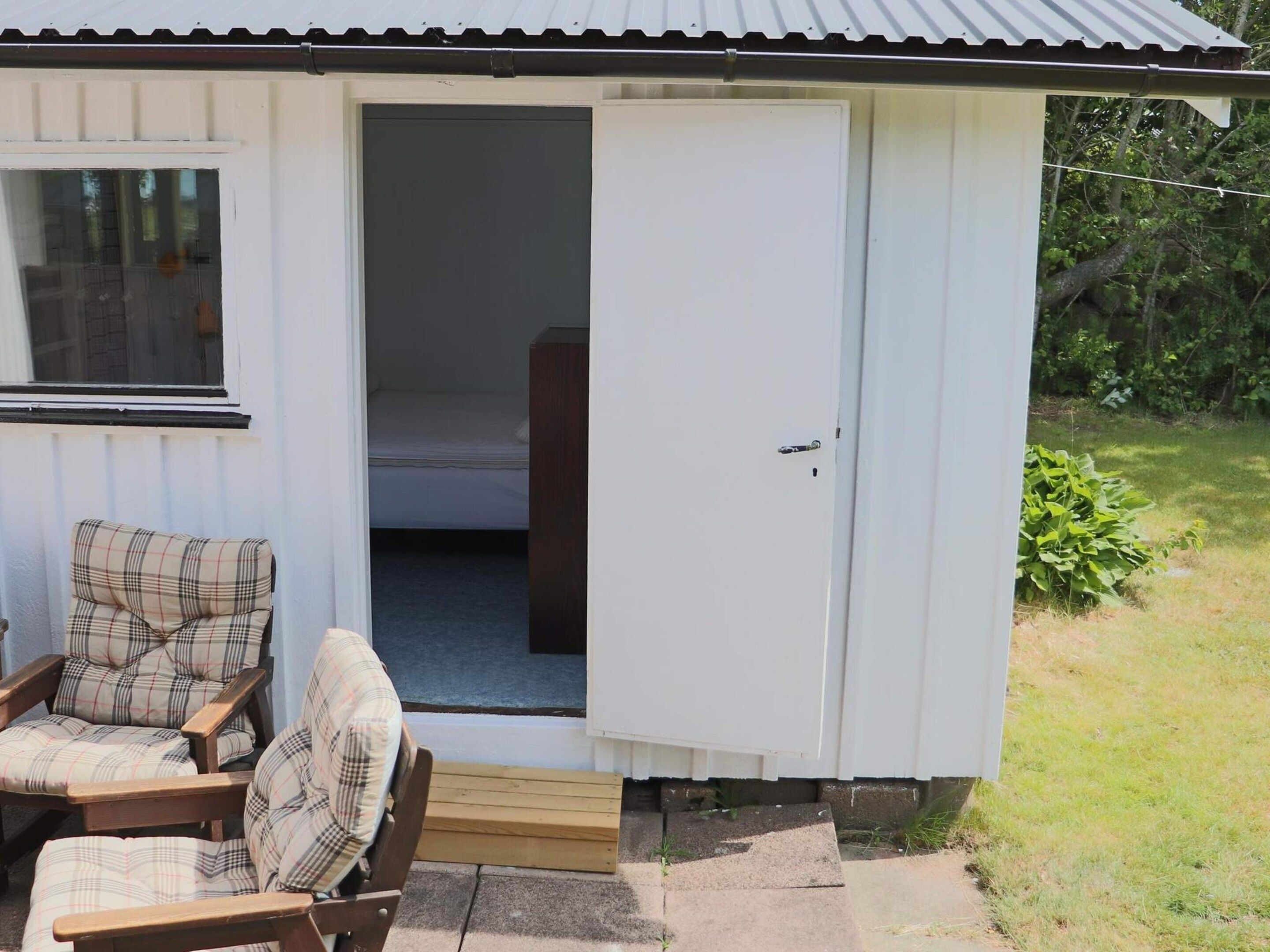 A small, single-room cabin with a door leading to a bedroom. There is a chair with a plaid cushion outside the cabin.