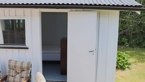 Eine kleine, einräumige Holzhütte mit einer Tür zu einem Schlafzimmer. Vor der Hütte steht ein Stuhl mit kariertem Sitzkissen.