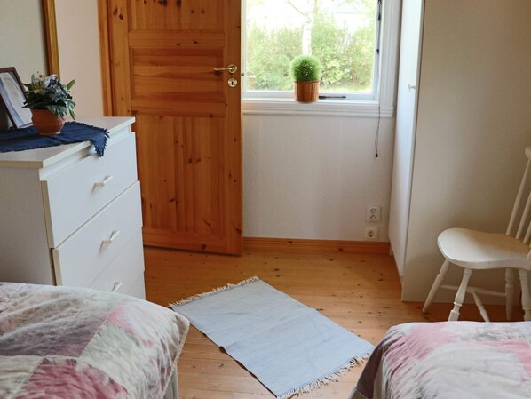 A bedroom with a wooden door, a white dresser, a chair, a bed with a patterned bedspread, and a small potted plant on the windowsill.