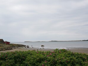 Paisaje costero con una playa arenosa, un edificio rojo y un promontorio rocoso al fondo.