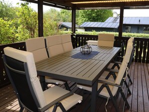 An outdoor patio with a wooden table and cushioned chairs, surrounded by a wooden deck and a view of a house with solar panels.