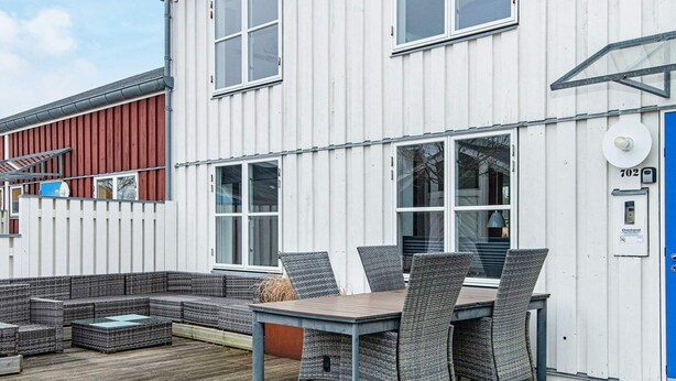 A modern outdoor seating area with a table and chairs, a wooden deck, and a white building with large windows and a blue door. - 8 Person Holiday Park Home in Ebeltoft (Ebeltoft)