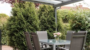 A covered patio with a glass table and four chairs, surrounded by dense greenery and a hedge. - Holiday Home Nispel 2 (Dornum)