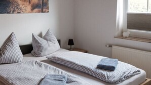 A double bed with checkered bedding, a nightstand with a lamp, a painting of a beach scene, and a window with a view of the outdoors. - Holiday Home Nispel 2 (Dornum)