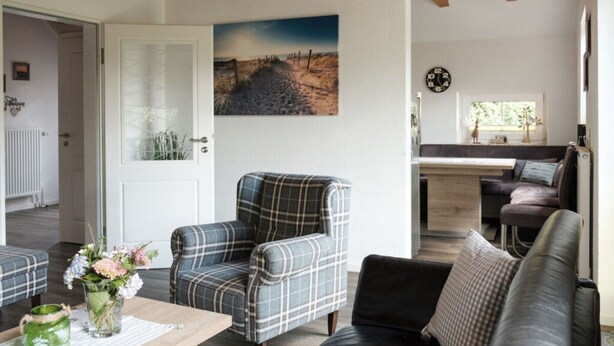 A cozy living room with a plaid armchair, a black sofa, and a wooden coffee table. A painting of a landscape hangs on the wall, and a clock is visible in the background. - Holiday Home Nispel 2 (Dornum)