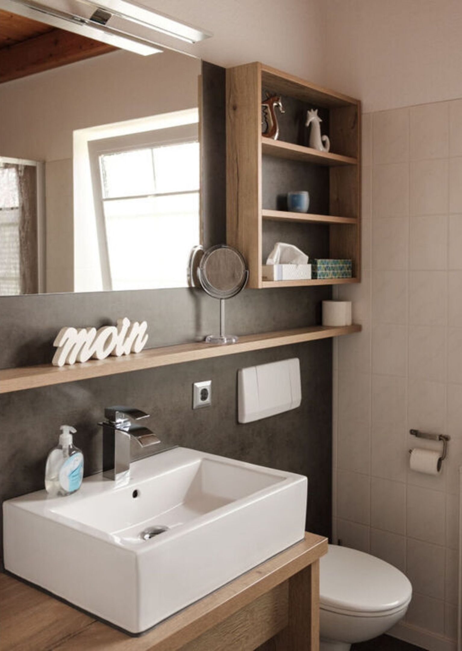 Et badeværelse med en hvid vask, et spejl, en træhylde med pynteting og et toilet.