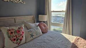 A bedroom with a bed, floral pillows, a decorative wall piece, a window with curtains, and a view of buildings. - Top floor condo in N. Topsail Beach. Views of the beach & Intracoastal Waterway! (North Topsail Beach)