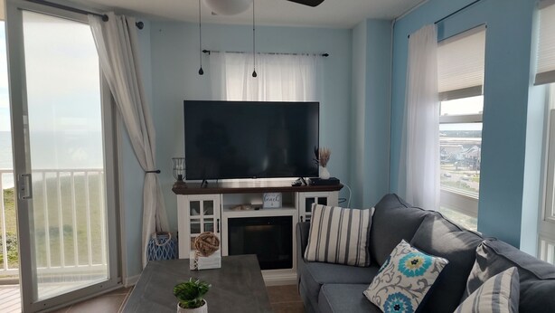 A living room with a flat-screen TV, a gray sofa with patterned cushions, a coffee table with a plant, and a view of the sea through large windows. - Top floor condo in N. Topsail Beach. Views of the beach & Intracoastal Waterway! (North Topsail Beach)