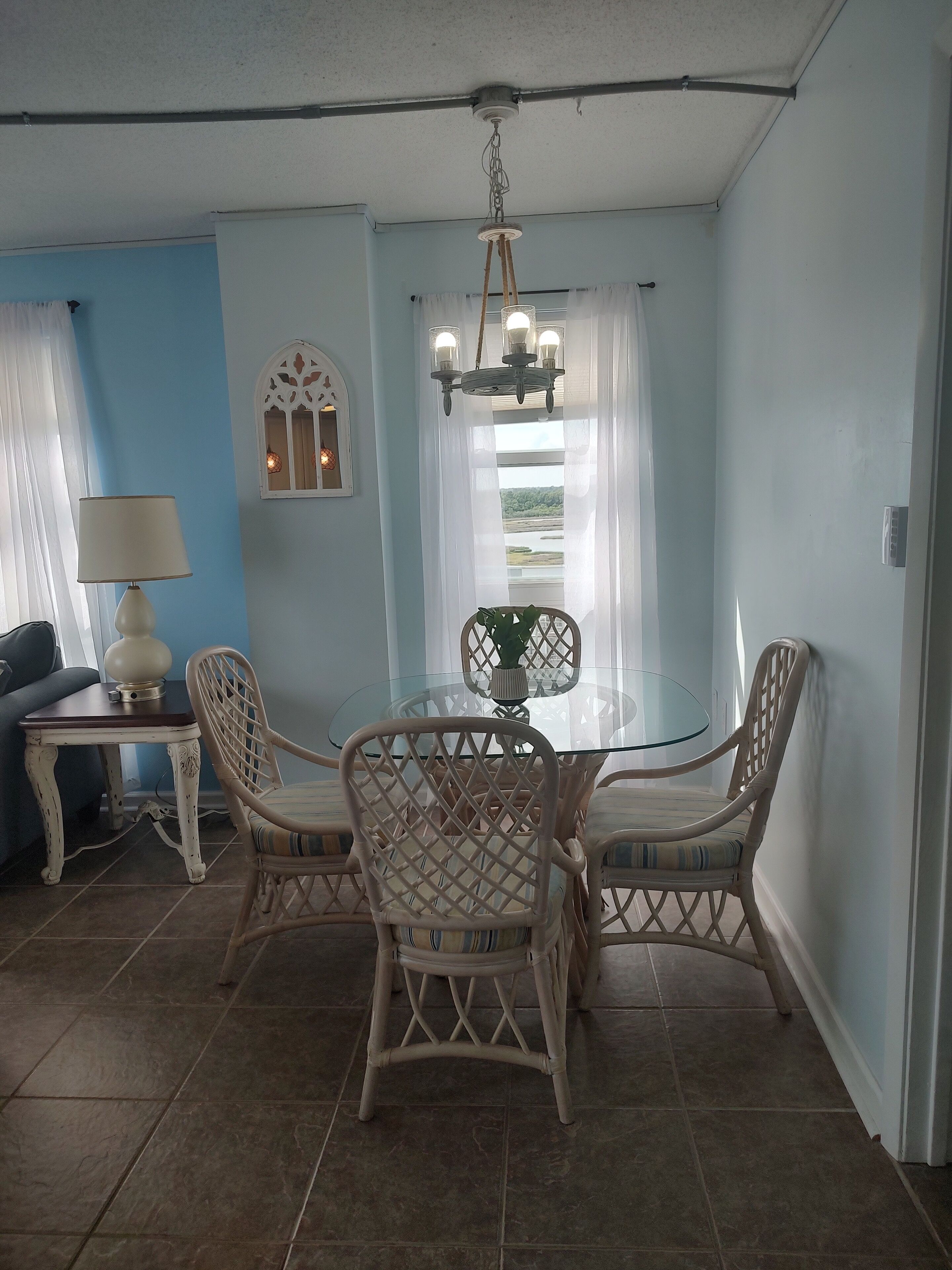 A dining area with a glass table and wicker chairs, a lamp, and a view through a window.