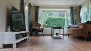 A modern living room with a large window, a white entertainment unit, a brown sofa, and a black chair. - Serene Forest Stay With Comfort (Putte)