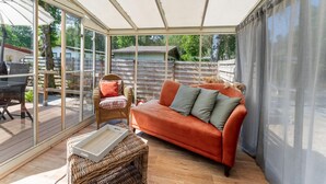 A sunroom with a glass enclosure, wicker furniture, and a red sofa. - Serene Forest Stay With Comfort (Putte)