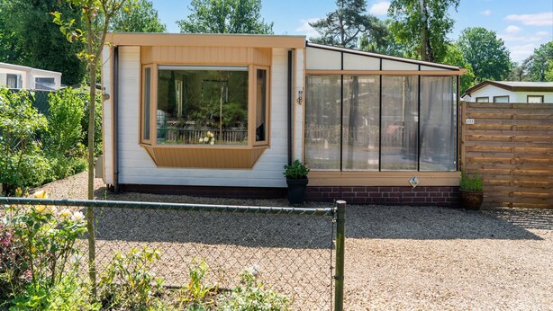A small, single-story house with a large window and a door, surrounded by a gravel driveway and a wooden fence. - Serene Forest Stay With Comfort (Putte)