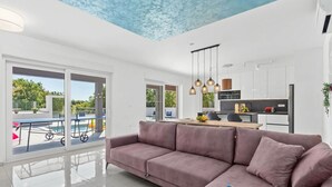 Una sala de estar moderna con un sofá grande, un comedor y una cocina. Hay ventanas amplias y vistas al exterior.