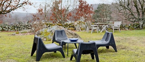 Un área de estar al aire libre con sillas de plástico y una mesa, rodeada por un muro de piedra y árboles.