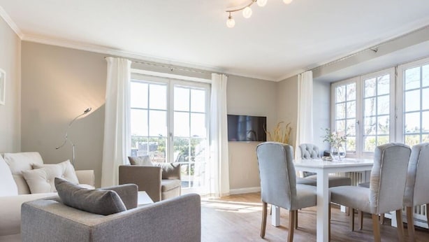 A modern living room with a sofa, chairs, a dining table, and large windows. - Holiday Home Hiirlonhus (Wenningstedt-Braderup)