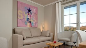 A modern living room with a beige sofa, a wooden coffee table, and a gray armchair. A colorful abstract painting is on the wall, and there is a large window with curtains. - Holiday Home Hiirlonhus (Wenningstedt-Braderup)