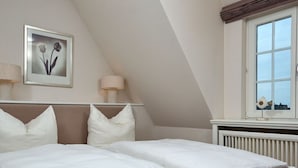 A bedroom with a large bed, two pillows, a framed picture on the wall, a window, and a radiator. - Holiday Home Hiirlonhus (Wenningstedt-Braderup)