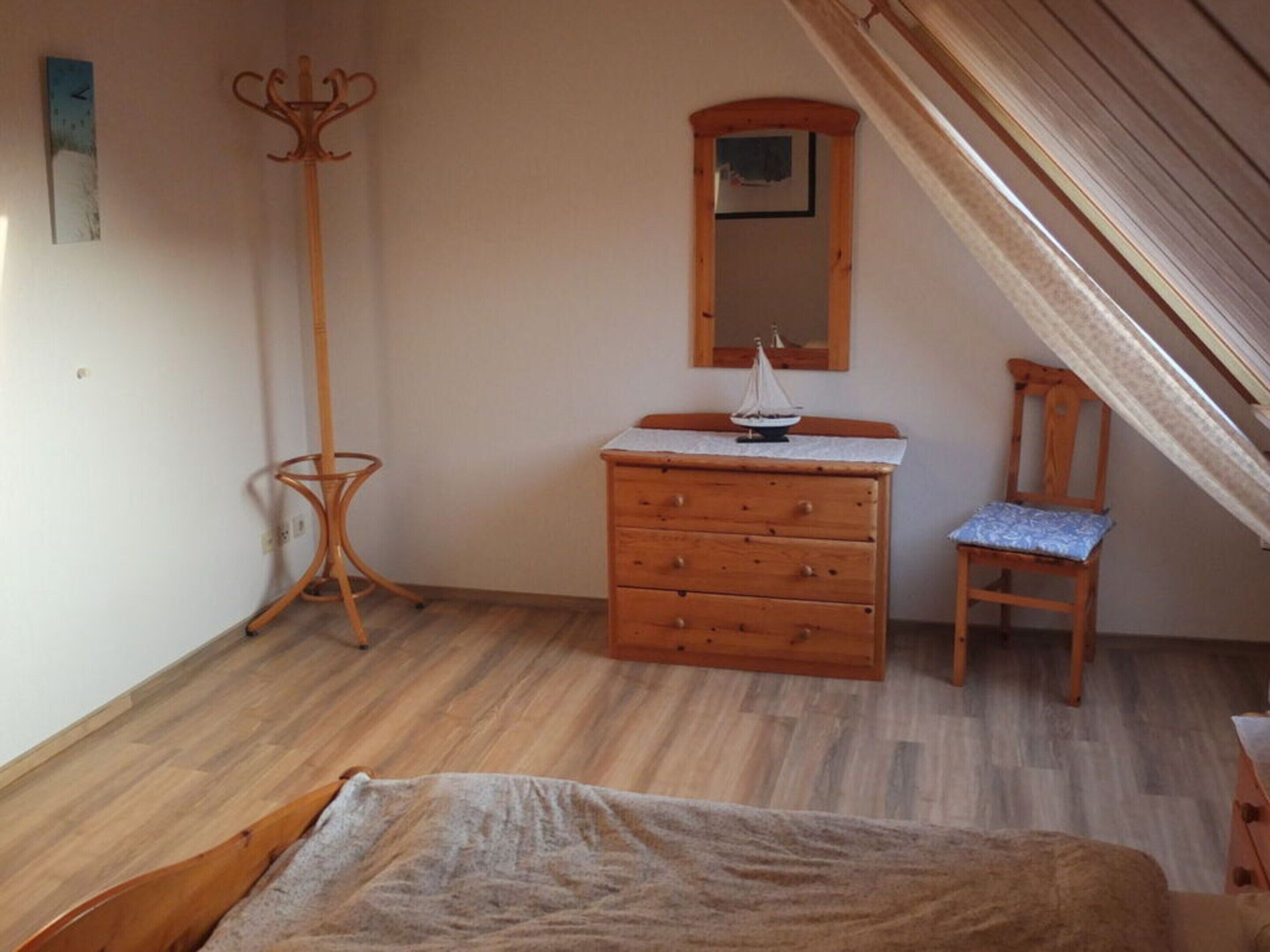 A bedroom with a wooden dresser, a chair, a bed, a coat rack, and a mirror on the wall.