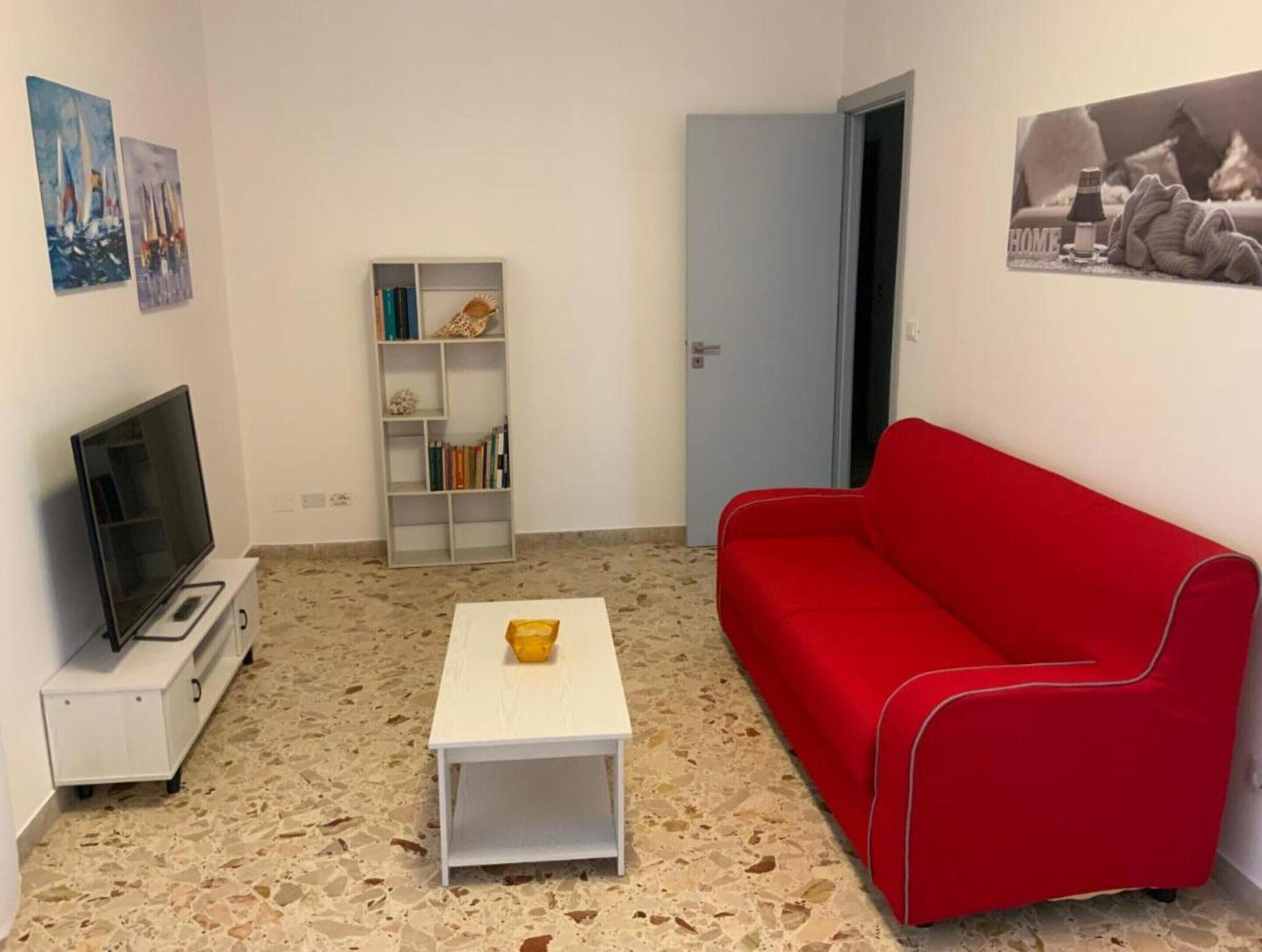 A living room with a red sofa, a white coffee table, a TV on a white stand, a bookshelf, and a door.