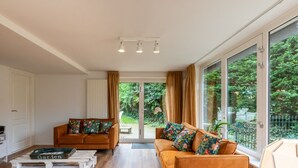 A spacious living area with two sofas, a coffee table, and a view of a garden through large windows. - Beautiful Bungalow With Garden (Biggekerke)