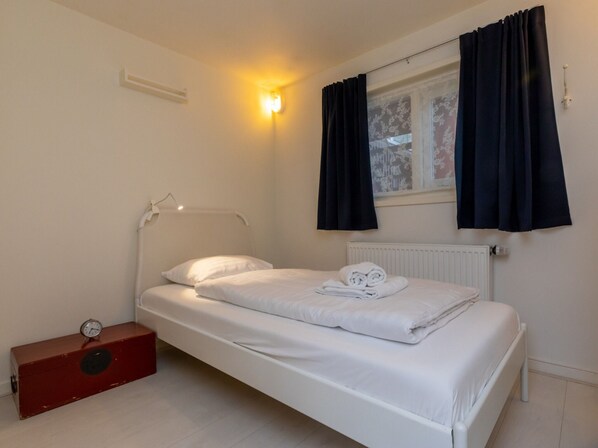 Una cama individual con ropa de cama blanca, una mesita de noche con un reloj y una ventana con cortinas oscuras.
