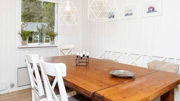 Un comedor con mesa de madera, sillas blancas, lámparas colgantes y una ventana con plantas.